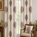 Off-white floral tree print curtain with pink flowers and grey leaves on a living room window, paired with a matching printed cushion on a wooden chair in an elegant Indian home.