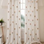 Elegant white sheer curtain with small orange floral prints, displayed on a large arched window in a warm minimalist room.
